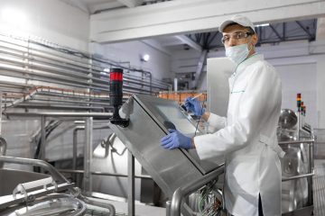 man in a white robe and a cap make a configure the digital screen technologist make set up of the production line while standing near digital screen at the department of dairy factory
